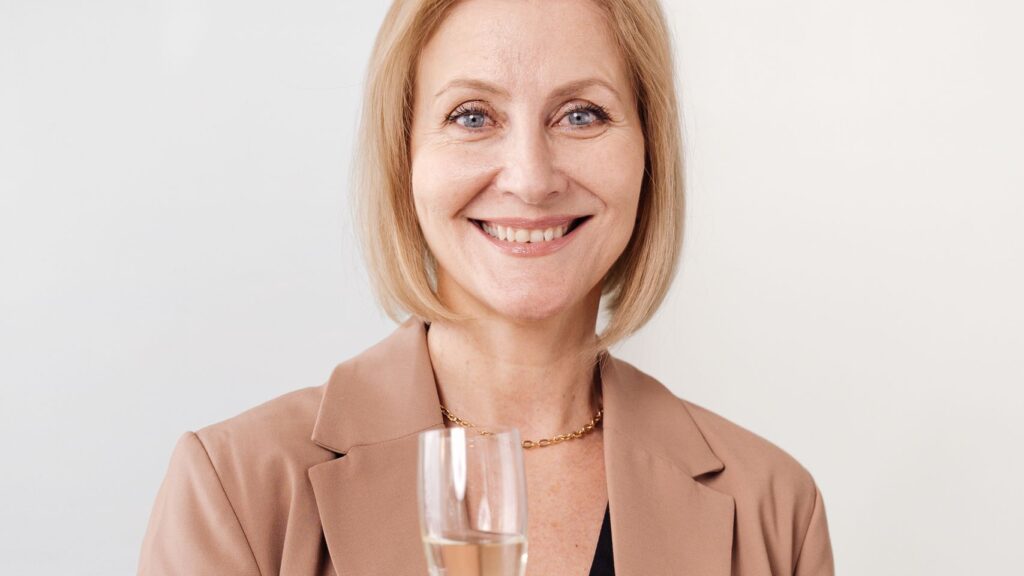 Elegant woman in a blazer holding champagne glass, smiling warmly indoors.